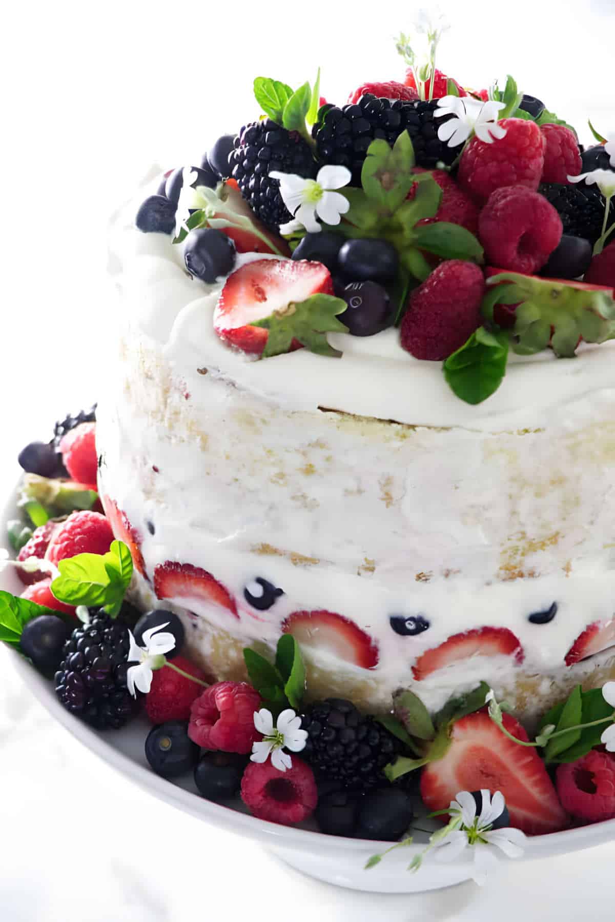 Chantilly cake on a pedestal strewn with fresh strawberries, blueberries, and raspberries.