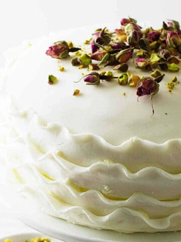 White layer cake on a cake pedestal with pistachios and rose buds on top.