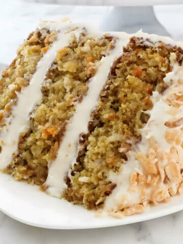 Slice of carrot cake on a plate with the cake on a pedestal in the background.